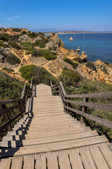A picture of a wooden path leading to Praia do Camilo in Lagos Portugal with a view of small rocky mountains and pacific ocean