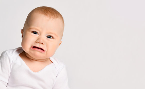 Chubby little kid in bodysuit, barefoot. He is about to cry, sitting on floor isolated on white studio background. Close up