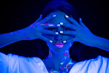young caucasian woman with fluorescent body art closing eyes with hands, look at camera through...