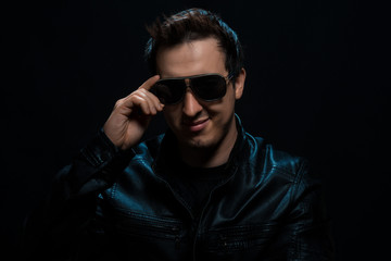 Young sexy stylish guy with glasses in a black leather jacket on a black background