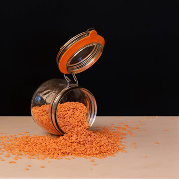 A glass kilner storage jar on its side with the lid wide open, red lentils, beans or pulses spill out on the a plain work surface.  The image is set against a plain black background.