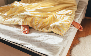 Fototapeta na wymiar Wake up in the morning. Men's legs under the blanket in the bed. Sunlight in a bedroom. Yellow striped wrinkled bed linen
