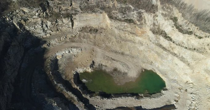 Aerial footage of a steep rock wall at a quarry. It shows mine terraces and water at the bottom of a mine