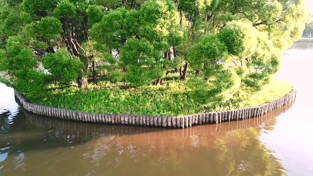 Aerial view of a big pond with island surrounded by calm water. Footage. Camera moving towards the green fresh tree leaves, vegetation on the roun decorative island.