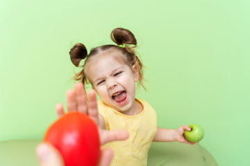 Fototapeta na wymiar a little girl of 4-5 years old holds an apple in her hand, the other hand extended forward to the tomato in a negative gesture. the refusal of children from vegetables in favor of fruits.