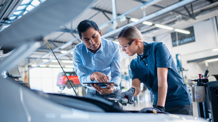 Instructor with a Tablet Computer is Giving a Task for a Future Mechanic. Female Student Inspects the Car Engine. Assistant is Checking the Cause of a Breakdown in the Vehicle in a Car Service. 