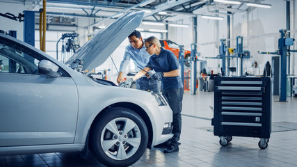 Manager Checks Data on a Tablet and Explains an Engine Breakdown to an Empowering Female Mechanic. Car Service Employees Inspect a Car with Internal Combustion Engine. Modern Clean Workshop.