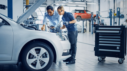 Manager Checks Diagnostics Results on a Tablet Computer and Explains an Engine Breakdown to an Female Mechanic. Car Service Employees Inspect Car's Engine Bay with a LED Lamp. Modern Clean Workshop.