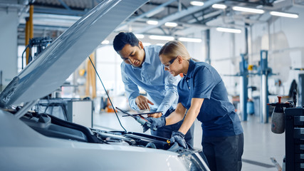 Instructor with a Tablet Computer is Giving a Task for a Future Mechanic. Female Student Inspects the Car Engine. Assistant is Checking the Cause of a Breakdown in the Vehicle in a Car Service. 