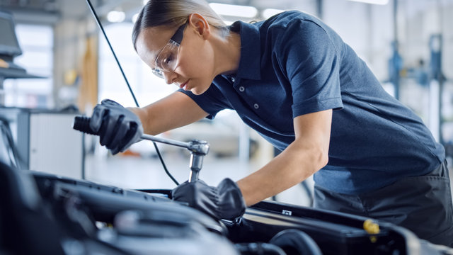 Beautiful Empowering Female Mechanic is Working on a Car in a Car Service. Woman in Safety Glasses is Fixing the Engine. She's Using a Ratchet. Modern Clean Workshop with Cars. 
