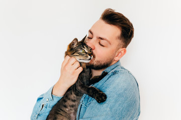 A happy young european man in blue denim shirt is holding and kissing  a cute brown tabby cat with...
