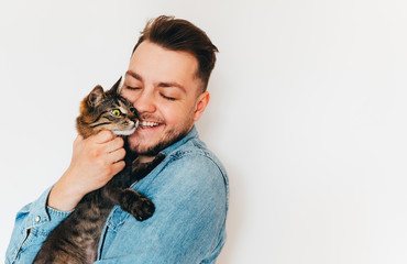 A happy smiling young man with beard in blue denim shirt is holding and hugging a cute brown tabby...
