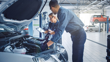 Two Mechanics in a Service are Inspecting a Car After They Got the Diagnostics Results. Female Specialist is Comparing the Data on a Tablet Computer. Repairman is Using a Ratchet to Repair the Faults.