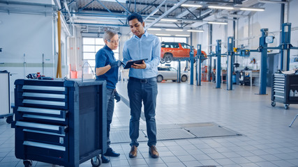 Car Service Manager and a Confident Female Mechanic Talk About Work Related Topics. They Use a Tablet Computer. Modern Clean Workshop with a Car on a Lift in the Background.