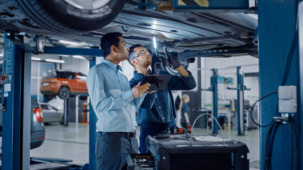 Manager Checks Data on a Tablet Computer and Explains the Breakdown to a Mechanic. Car Service Employees Inspect the Bottom of the Car with a LED Lamp. Modern Clean Workshop.