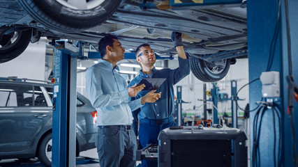 Manager Checks Data on a Tablet Computer and Explains the Breakdown to a Mechanic. Car Service Employees Inspect the Bottom and Skid Plates of the Car. Modern Clean Workshop.