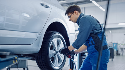 Plakat Mechanic in Blue Overalls is Unscrewing Lug Nuts with a Pneumatic Impact Wrench. Repairman Works in a Modern Clean Car Service. Specialists Removes the Wheel in Order to Fix a Component on a Vehicle.