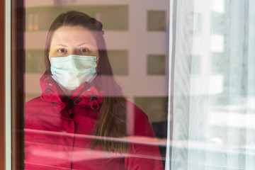 Portrait of a girl or young woman in a medical mask being quarantined at home during the coronavirus pandemic. Self-isolation, view through glass, walking on the balcony of an apartment in city