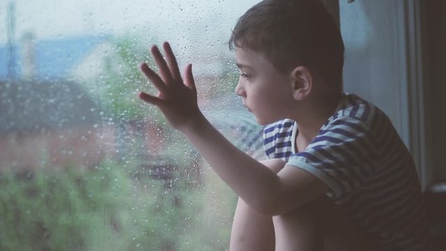 a little boy sits outside a rainy window and is sad. A window in rainy weather behind which a small sad boy sits, a child is hard going through self-isolation, orphaned children in orphanages and boar