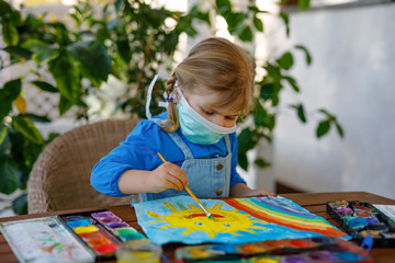 little toddler girl in medical mask painting rainbow with water colors during pandemic coronavirus quarantine disease. Children painting rainbows around the world with the words Let's all be well.