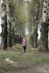 Obraz premium A girl walks along a spring poplar alley. It goes between two rows of tall trees. Dressed in a spring jacket, a scarf around her neck. Around the blossoming greens on the trees.
