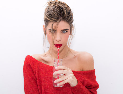 chica rubia bebiendo con pajita con jersey y pintalabios rojo en fondo blanco mirando de frente