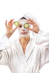 A beautiful young woman with white cream on her face and juicy fresh cucumbers in her eyes looks at the camera. White isolated background.