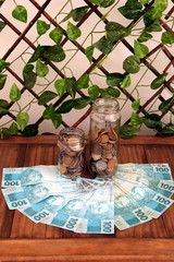 Brazilian money with several 100 reais bills and glass jars of different sizes with coins on wooden table