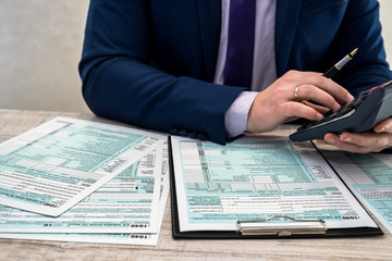 A man in a business suit writes a 1040 tax form in the office. Male hands fill on paper with calculator at workplace