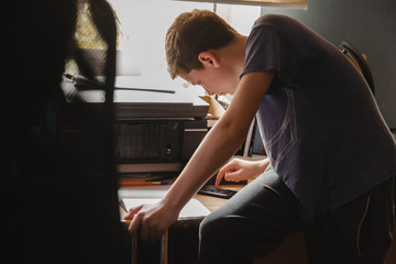 A teenage boy in home clothes at home sits on a table and learns lessons using a smartphone