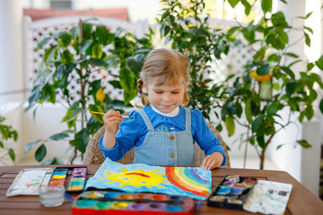 little toddler girl painting rainbow and sun with water colors during pandemic coronavirus quarantine disease. Children painting rainbows around the world with the words Let's all be well. Happy child