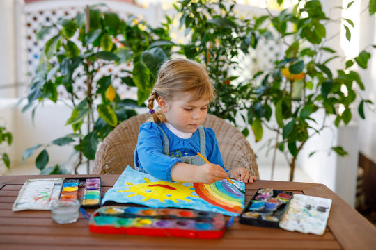 Little Toddler Girl Painting Rainbow And Sun With Water Colors During Pandemic Coronavirus Quarantine Disease. Children Painting Rainbows Around The World With The Words Let's All Be Well. Happy Child