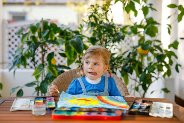 little toddler girl painting rainbow and sun with water colors during pandemic coronavirus quarantine disease. Children painting rainbows around the world with the words Let's all be well. Happy child