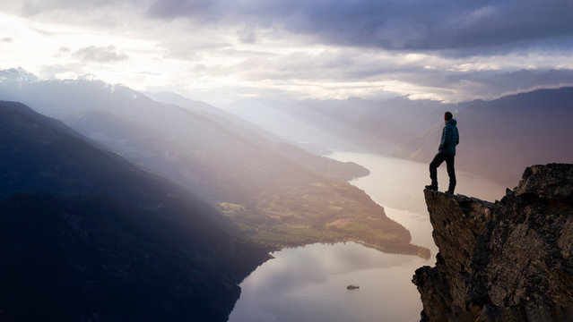 Fantasy Adventure Composite with a Man on top of a Mountain Cliff with Dramatic Landscape in Background during Sunset or Sunrise. Landscape from British Columbia, Canada.