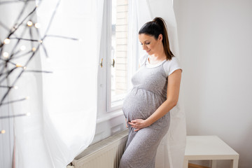 A pregnant woman standing next to the window with hands on her b