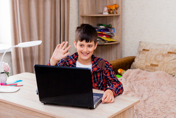 The boy sits at a table with a laptop and does homework. Side view. boy talking on skype