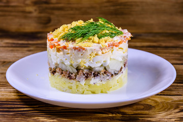 Plate with traditional russian salad Mimosa on a wooden table. Layered salad with potatoes, sardine, cheese, carrot, eggs and mayonnaise