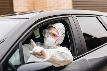 Man in protective suit, medical mask and rubber gloves for protect from bacteria and virus is sitting in a car and driving to hospital. Concept protection during the covid 19 coronavirus pandemic.