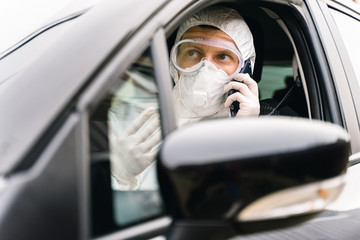 Man in protective suit, medical mask and rubber gloves for protect from bacteria and virus is sitting in a car and talking on the phone. Concept protection during the covid 19 coronavirus pandemic.