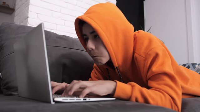 Adult teenager in orange sweater plays on laptop at sofa at home