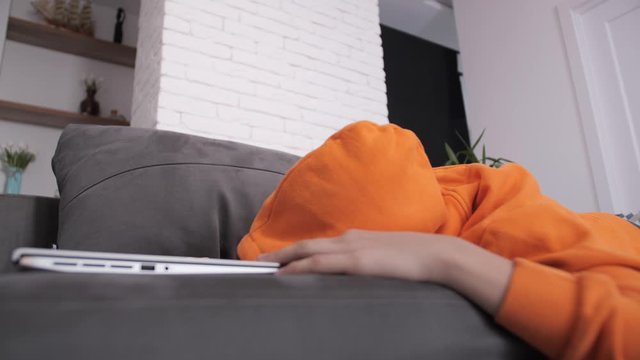 Adult teenager in orange sweater plays on laptop at sofa at home
