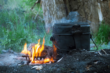 Hiking smoked pot on the coals, old black military bowler in campfire in summer evening. Selective focus.