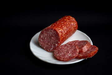 Slices of tasty delicious pepperoni sausage on white plate on dark black background