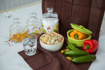 Decorated with lettuce, side view, selective focus . Orange pepper in a green bowl . Water in a glass. Brown tablecloth. Salad with ham, bell pepper, mushrooms, pickles, white sauce, on a white plate.