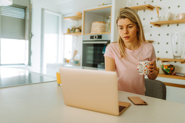 Portrait of young blonde woman drinking coffee and using laptop early in the morning. Young woman is working from home, because she is in self quarantine.