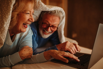 Close photo of good looking elderly couple using laptop in bed covered with blanket.