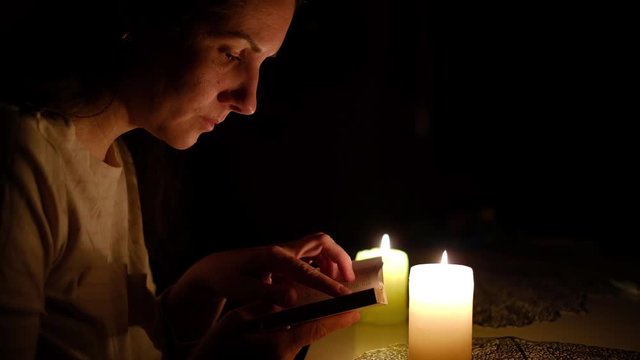 A woman sits and reads a bible verse by candlelight at night, watches the text with her finger, close-up