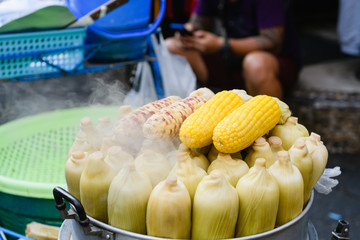 Raw, steamed and grilled sweet yellow corn cobs in big pan at asian thai street food market close up