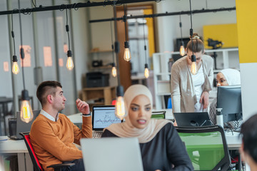Fototapeta na wymiar African muslim girl wearing hijab working on a laptop in a modern startup company. While being gossiped by her european coworkers.