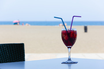 Sangria glass with fruit pieces and plastic straws on beach terrace. Refreshing alcoholic drink on table with sea views on background. Summer holidays leisure time, tempting happy hour concepts